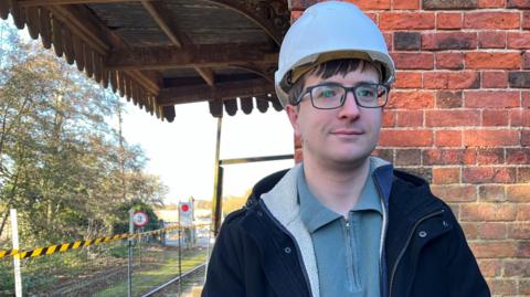 Alex Bramer from The Middleton Towers Restoration Group stands in front of a red brick wall. He is wearing a white hard hat, dark blue coat and light blue fleece underneath. 