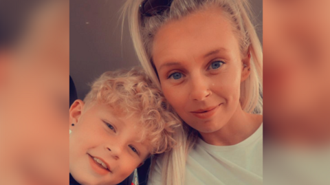 A woman and her son are smiling into the camera. The woman has long blonde hair, past her shoulders, while her son has shorter, curly hair.