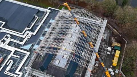 A yellow crane is extended over the roof of The Sands Centre. white metal rigging appears to have been constructed over the building's roof.