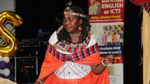 Bernadetta Omondi representing the Black History Month committee in Peterborough at a previous event. She wears a white top with brightly coloured orange fabrics wrapped around her arms and waist. She also wears a brightly coloured head dress and jewellery around her neck. 