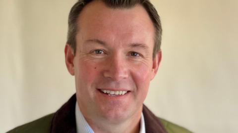 Man with dark brown hair posing for a photo with a cream wall behind him, wearing a pale blue shirt and green jacket with brown collar.