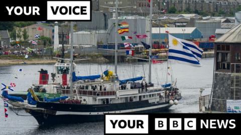 A tall ship leaves Aberdeen Harbour during the Parade of Sail. There are a number of flags on the boat, including a large Uruguay flag. There are warehouses visible in the background, as well as crowds on the harbourside. 