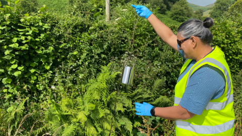 Susie Roy testing water. She stands in front of foliage including ferns, holding a string attached to a canister while wearing latex gloves. She has grey and dark hair pulled back in a bun and a blue-striped shirt under a hi-vis jacket