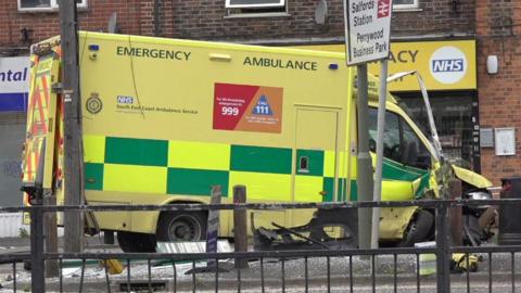 An ambulance which has crashed into a bus stop. The image shows the right side of the ambulance. Its front wing and wheel are badly damaged