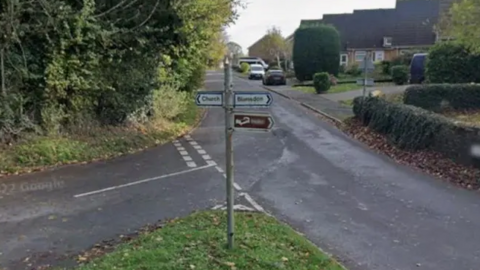 GoogleMaps image of a road and crossroads with one sign pointing to 'Blunsdon'. There are grass verges on both side of the road and trees lining the left-hand side