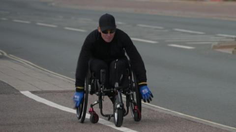 Wheelchair athlete Lexi Chambers training for her challenge along the seafront in Exmouth. She is wearing an all black outfit, with black glasses and a black cap. She is wearing blue gloves. 