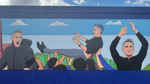 Part of a mural installed opposite Portman Road football stadium in Ipswich. This section of mural details three scenes and figures of Kieran McKenna, the former Ipswich Town FC manager.