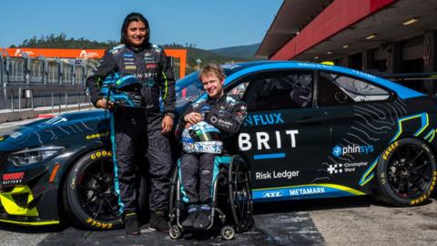 Two people in Team Brit race suits in front of a BMW M4 GT4 Race Car. It is black with blue and yellow accents. They are at the Circuit de Spa-Francorchamps in Belgium.