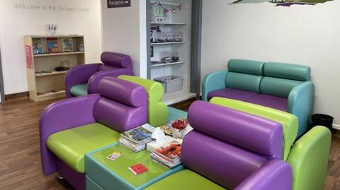 The waiting room in the Orchard Centre has five green and purple armchairs and a sofa along with a table with magazines and pens. There are two bookshelves lining the walls and writing on the wall that reads 'Welcome to the Orchard Centre'.