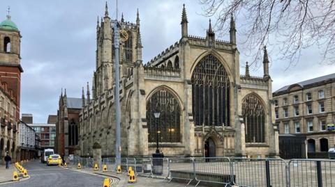 A view of the minster from the front from the left.  The church has three large arched windows to the front and a raised middle section.  A tower rises at the back of the church.  The church is built from a cream stone.