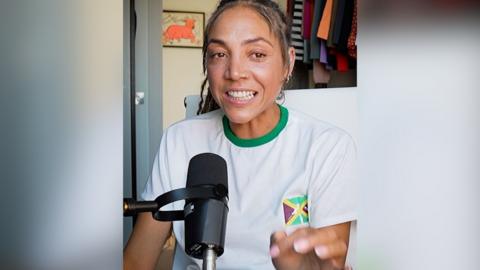 A woman, Miquita Oliver, wears a white T-shirt with the Jamaica flag and has long brown hair. She sits behind a microphone