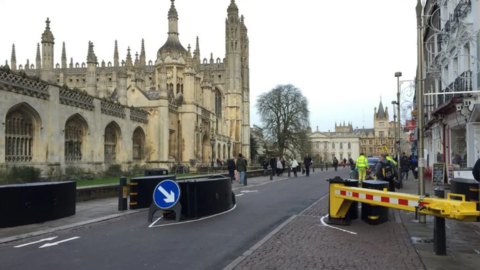 A pedestrianised street before the large chapel of King's College Cambridge. There are barriers at the beginning of the road, and pedestrians walking on pavements. 