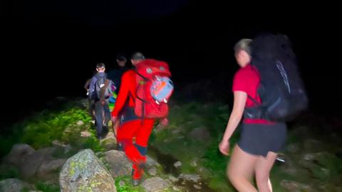 Shot of three people walking down a mountain in the dark. The shot is blurry. The volunteers are wearing red and have torches. The ground is mossy.