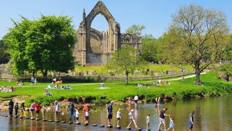 People walking across stepping stones on the Bolton Abbey estate, with abbey ruins in the background