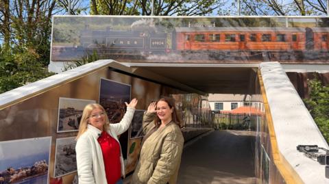A lady on the left in a red jumper wearing a white coat, she has glasses on with blonde hair. The lady to her right has brown hair and is wearing a green coat. They're stood under a newly decorated viaduct pointing to an image of a steam train which used to go to Hornsea. 