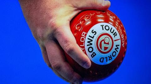 Fingers gripping a red bowl which is marked with the words "world tour bowls". The background is blue.