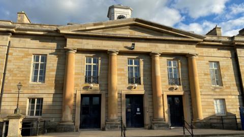 A two-storey Georgian style court building made form yellow stone with long elegant windows and four columns around three black doors. On the roof there is a white clock tower
