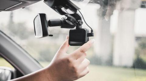 Video recording equipment in a car that has been set up on the rear-view mirror.  The photograph is a close-up of the driver's hand adjusting the kit. The view outside the car is blurred.