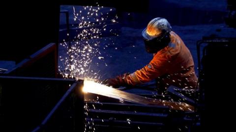 A steelworker in full protective gear working at a steel plant. Sparks are flying as he appears to cut through a material