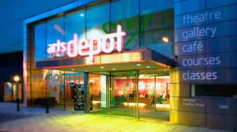 Foyer of a building lit with different coloured lights with the words arts depot in pink and a list of things inside written on the wall including theatre, gallery, cafe, courses and classes