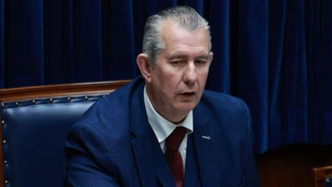 Edwin Poots has short grey hair and is wearing a blue blazer, red tie and white shirt.