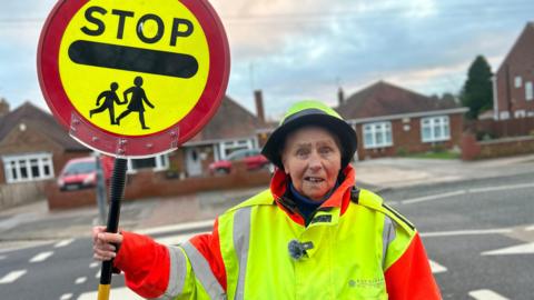 An elderly lady wearing a high-vis uniform holding her lollipop next to the zebra crossing she has worked on for 40 years