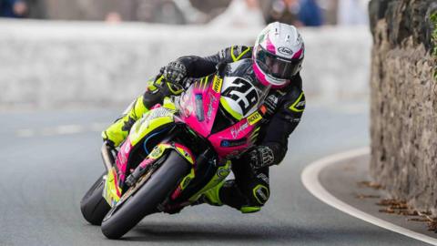 Naughton rounding a bend with a stone wall on the left on the Billown Course. His leathers are black and yellow, his helmet is white and he is on a pink and yellow bike.