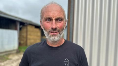 A man with grey hair and a grey beard, wearing a navy top. he is standing in front of a farm building