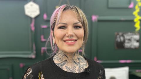 Naomi Dalby has pink and blonde hair, a septum and nose piercings, and tattoos on her neck. She is smiling at the camera and wearing a black top.