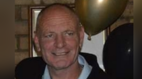 Close up of Gary Hindle in a white shirt and dark jacket. He is standing in front of a picture frame and has a gold and a black balloon by his head.