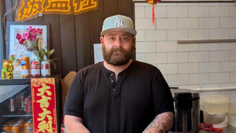 John Leedham looks into camera. He wears a black shirt and Yankees cap and stands in a kitchen area with containers, bottles, and utensils on the counter. A fridge with jars and flowers sits beside a neon sign and a red lantern hangs from the ceiling.
