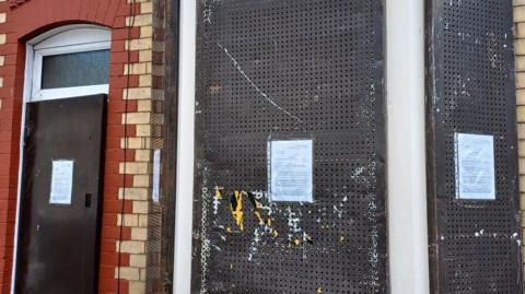 The front window of a brick terrace house has been boarded off with sheets of metal. A4-sized notices have been stuck to the window and front door. 