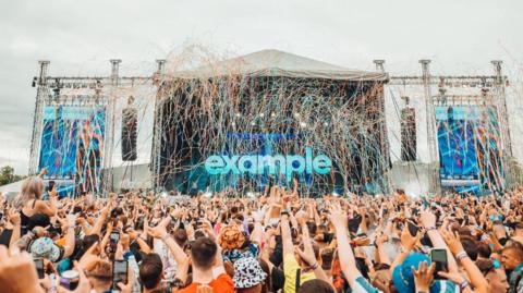 A crowd of people standing outside surrounding a stage, with confetti hanging above their heads.