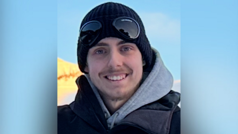 A photograph of Liam Derrett, a young white man wearing a thermal coat with a grey hoody underneath. He is wearing a woolly hat with snowboarding goggles over it. 