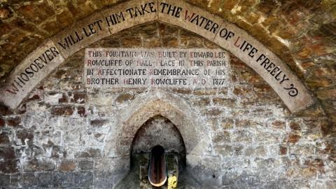 A fountain coming out of a stone wall. The wall has faded inscriptions on it.