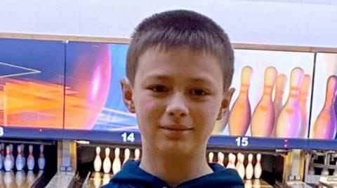 A close-up photo of a boy looking straight at the camera. He has short brown hair and he is wearing a blue hoodie. He is in a ten-pin bowling alley, with lanes and sets of pins visible behind him.
