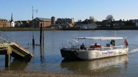 A white boat on the River Ouse. On the side of the boat it says "Lynn Ferry" in blue letters. There are two passengers and a pilot on board.