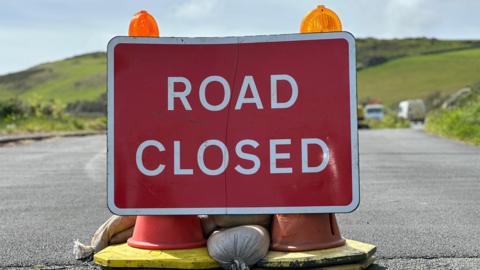 A road closed sign on Marine Drive with the road snaking off behind and green hills above.