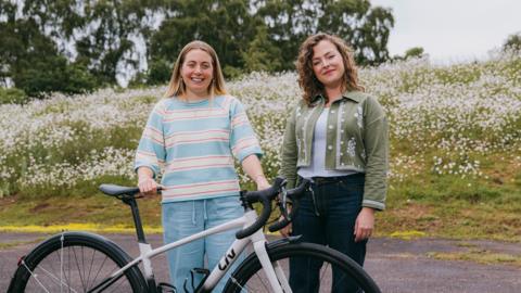 Two women standing behind a bicycle in a field of wild flowers