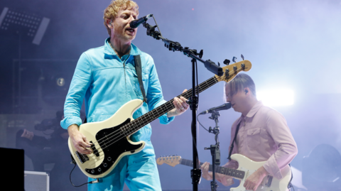 Two band members playing guitars and singing into microphones on stage at Reading Festival.  One is wearing a bright blue tracksuit jacket and trousers, the other is wearing a pale pink shirt.