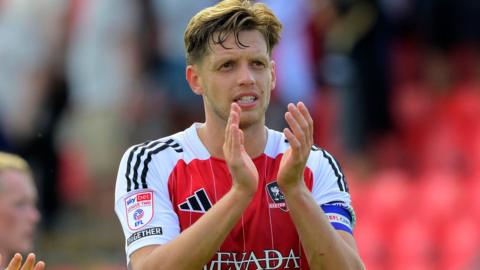 Exeter City defender Jack Fitzwater claps after having scored one of the goals in a 2-0 win over Accrington Stanley. He has short brown hair and is wearing the red and white home shirt of Exeter. 