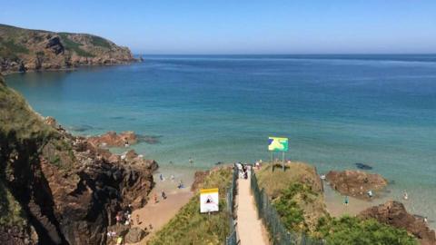 A view of a path leading to a turquoise sea with cliffs in the background.