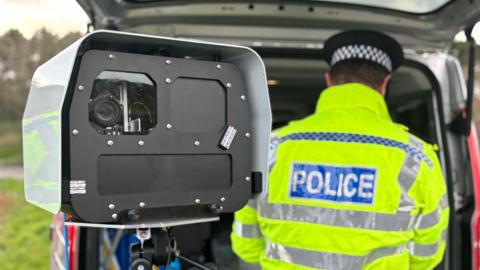 A large rectangular speed camera on a tripod is visible. Behind it, a police officer in a high-visibility jacket is working in a police van. 