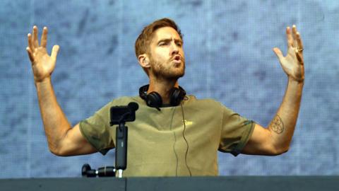 Calvin Harris holds his hands aloft while wearing a set of headphones on stage. 
