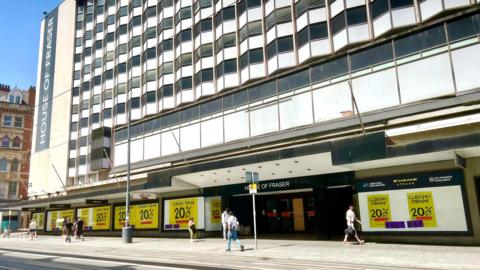 A photo of a high street department shop with 20 percent off signs in the windows with yellow. There are people walking in front of the shop. 