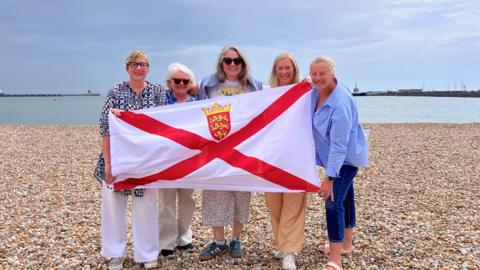A picture of the five women standing on a beach with a flat sea behind them. They are holding the Jersey flag.