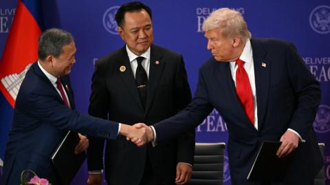 Thailand's Prime Minister Anutin Charnvirakul (C) watches as Cambodia's Prime Minister Hun Manet (L) and US President Donald Trump (R) shake hands. All three are wearing dark suits. Trump has a red tie, Anutin has a dark tie and Manet has a dark red tie. They are standing against a blue background with "delivering peace" written repeatedly on it.