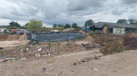 Mounds of waste, including tyres, lie on a bare patch of ground next to a large barn.