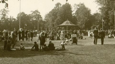 A black and white photo of people in a park. There is a bandstand in the distance with a conical roof. Some people are sitting on the grass while others are walking around. There is a large group of people near the bandstand. There are trees in the background.