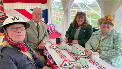 Four people sitting around a table wearing 1940s clothing. They are smiling to the camera and on the table are flags of Great Britain. They are sitting inside a marquee.
